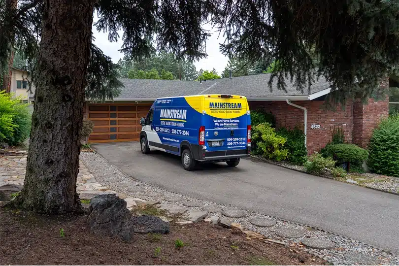 The Mainstream van parked in a driveway of a brick home with a giant fir tree in the foreground