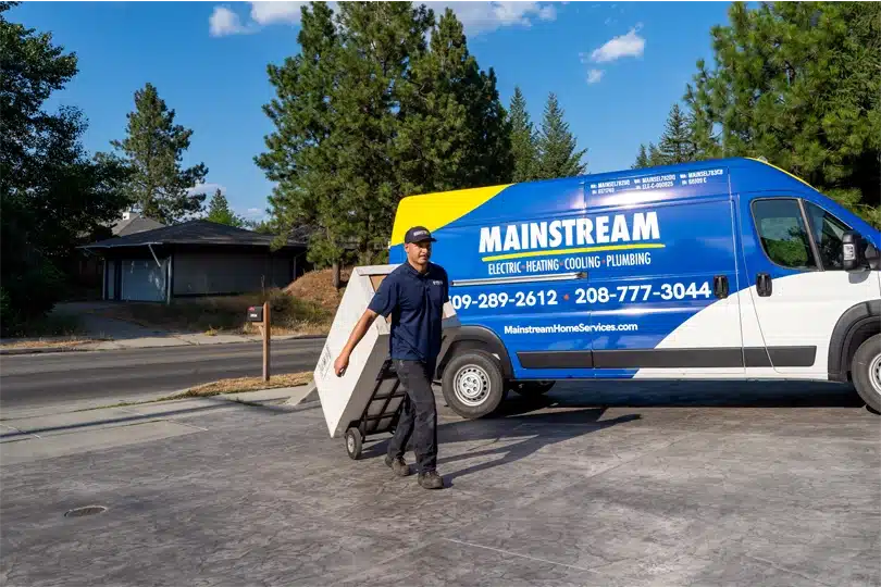 A Mainstream plumber walks up a driveway to deliver a new water heater to a home