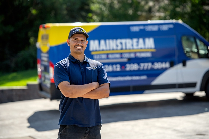 A Mainstream Electric, Heating, Cooling, & Plumbing technician stands with his arms crossed in front of the Mainstream work van in the background parked on a driveway